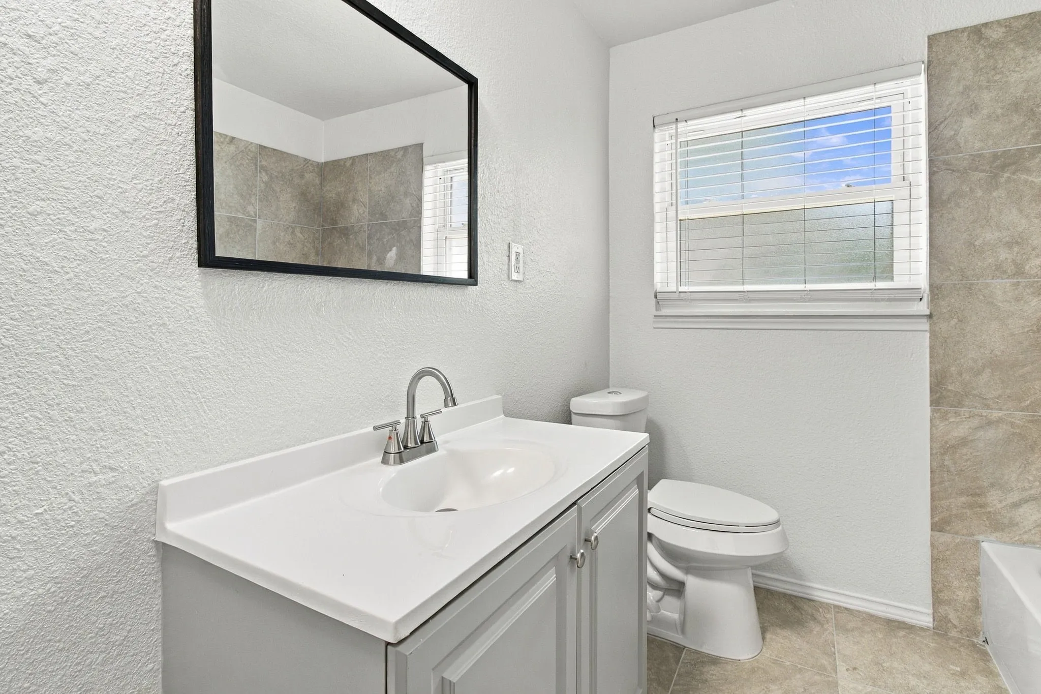 White vanity with integrated sink, rectangular mirror, brushed nickel faucet, tiled flooring, and a window with blinds