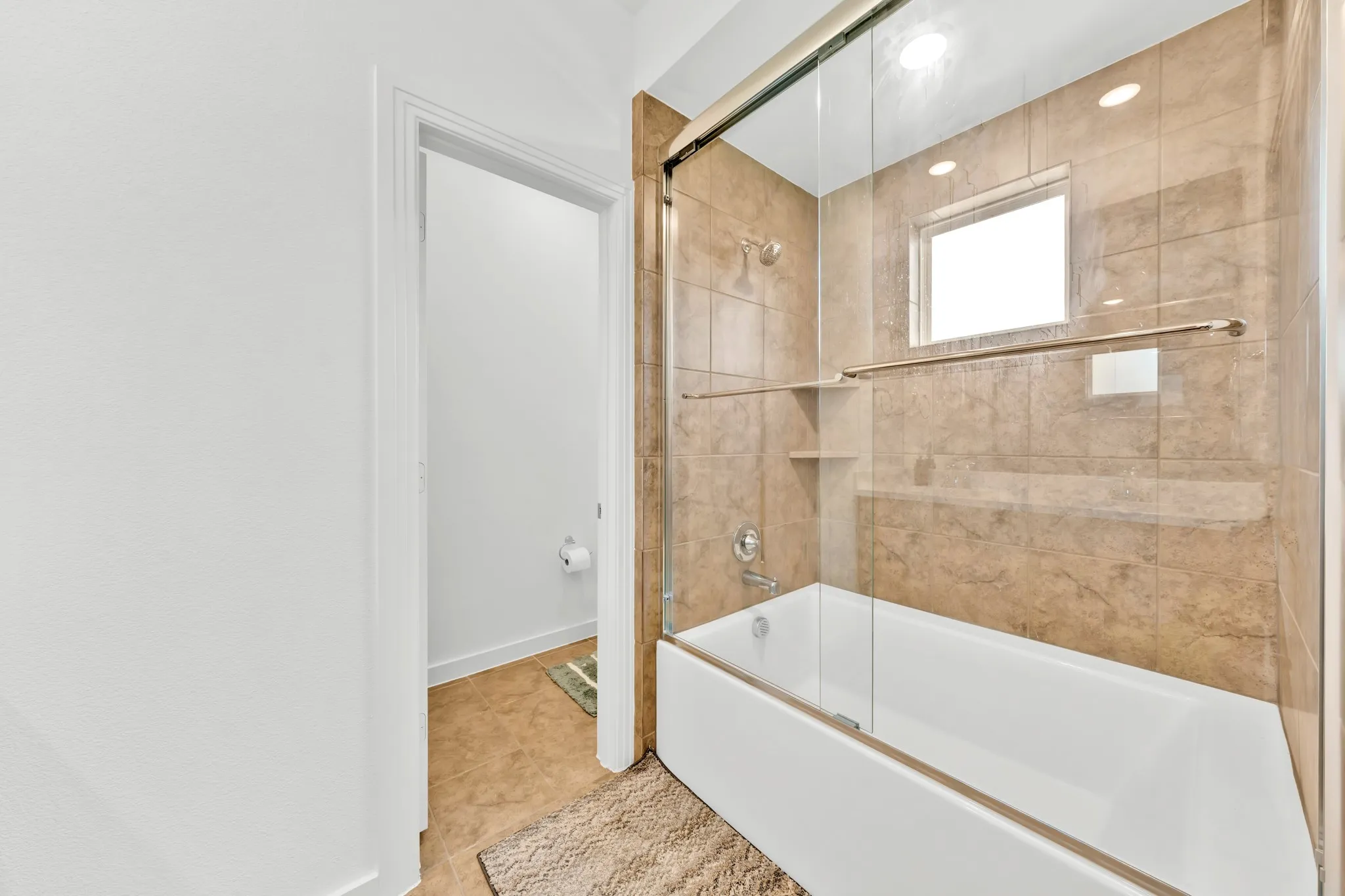 White bathtub with a sliding glass enclosure, stone-finish tile surround, built-in shower niche, and a frosted window