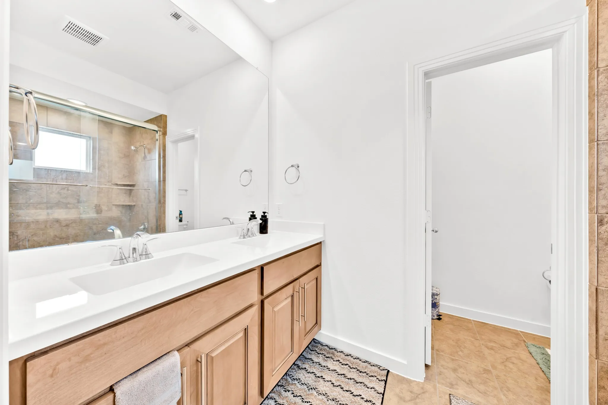 Double vanity with integrated sink, wood-finish cabinetry, frameless mirror, and glass-enclosed shower with tile surround