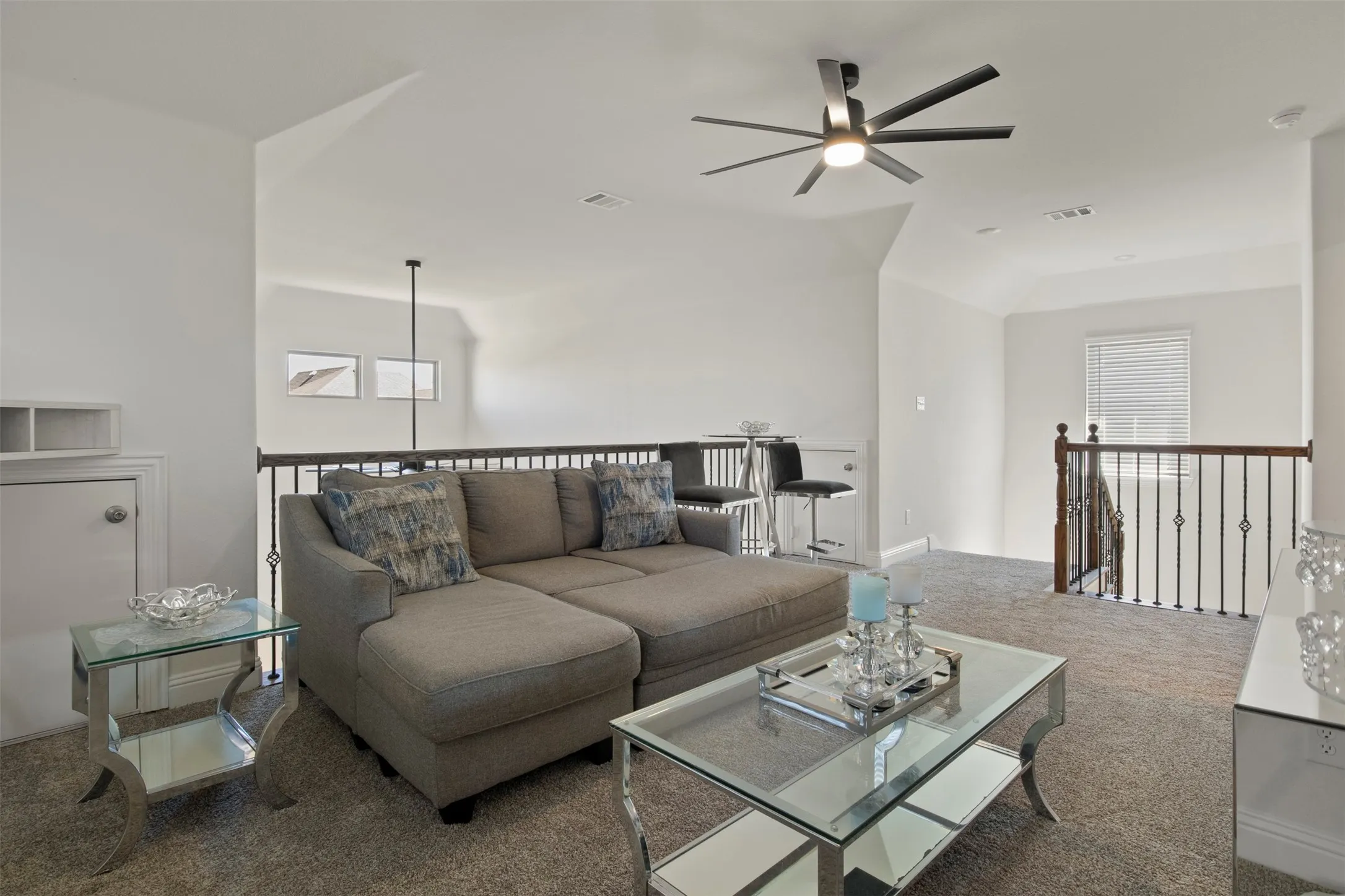 Carpeted loft area featuring a ceiling fan, wrought iron railing, and natural light from a window with blinds