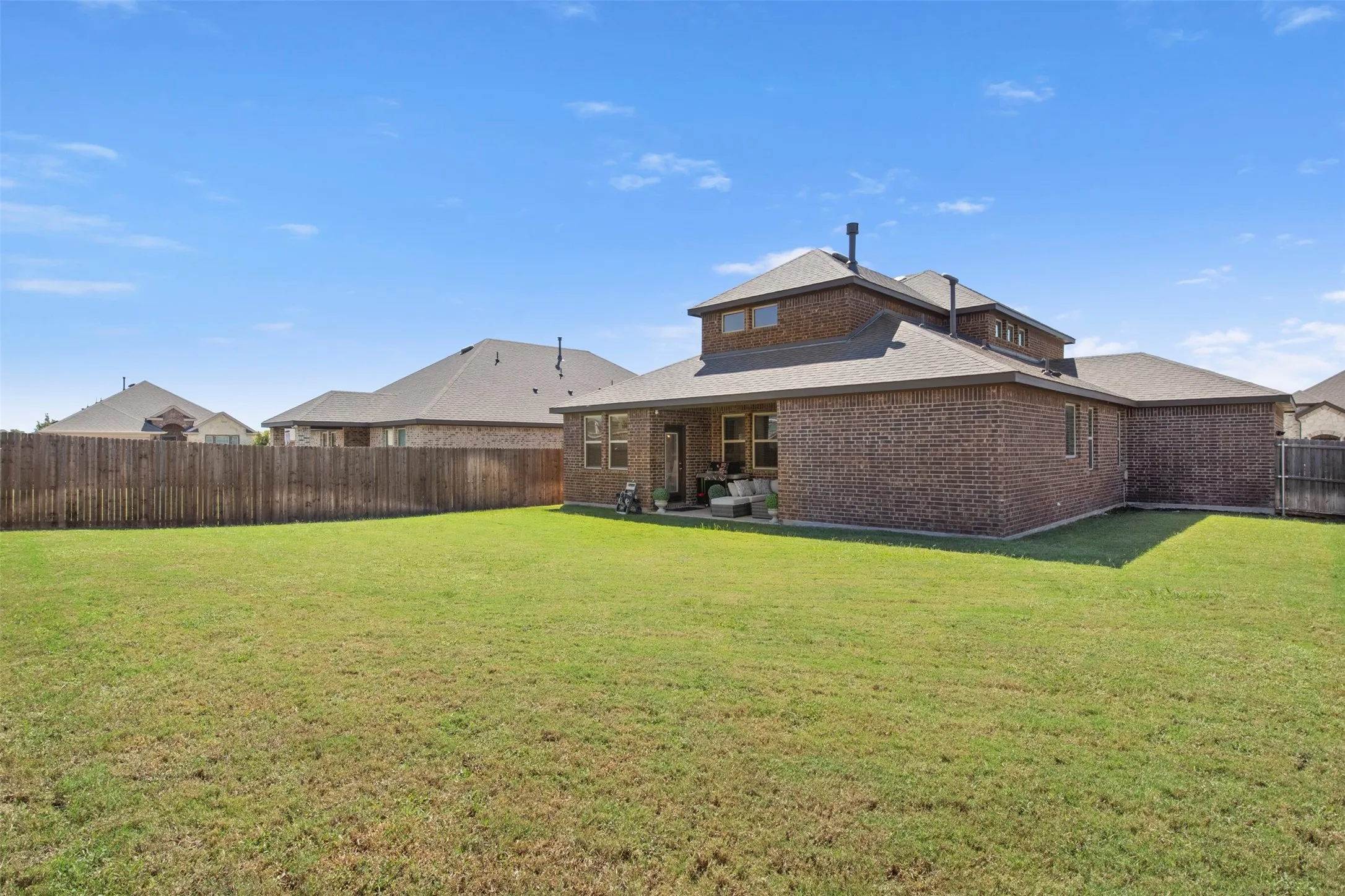 Expansive grassy yard with a wooden privacy fence, a brick exterior, a covered patio, and multiple windows