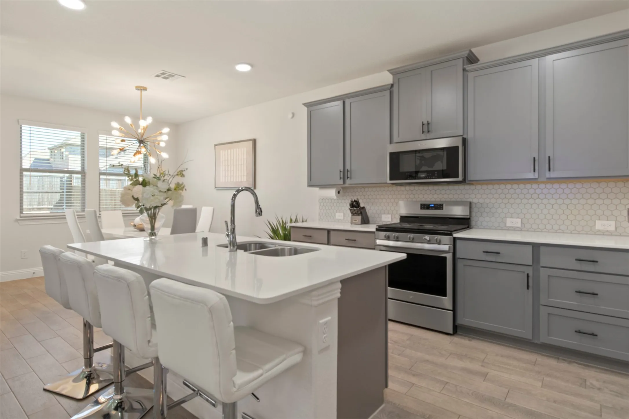 Kitchen featuring an island with a built-in sink, gray cabinetry, white countertops, stainless steel appliances, and a white hexagonal tile backsplash