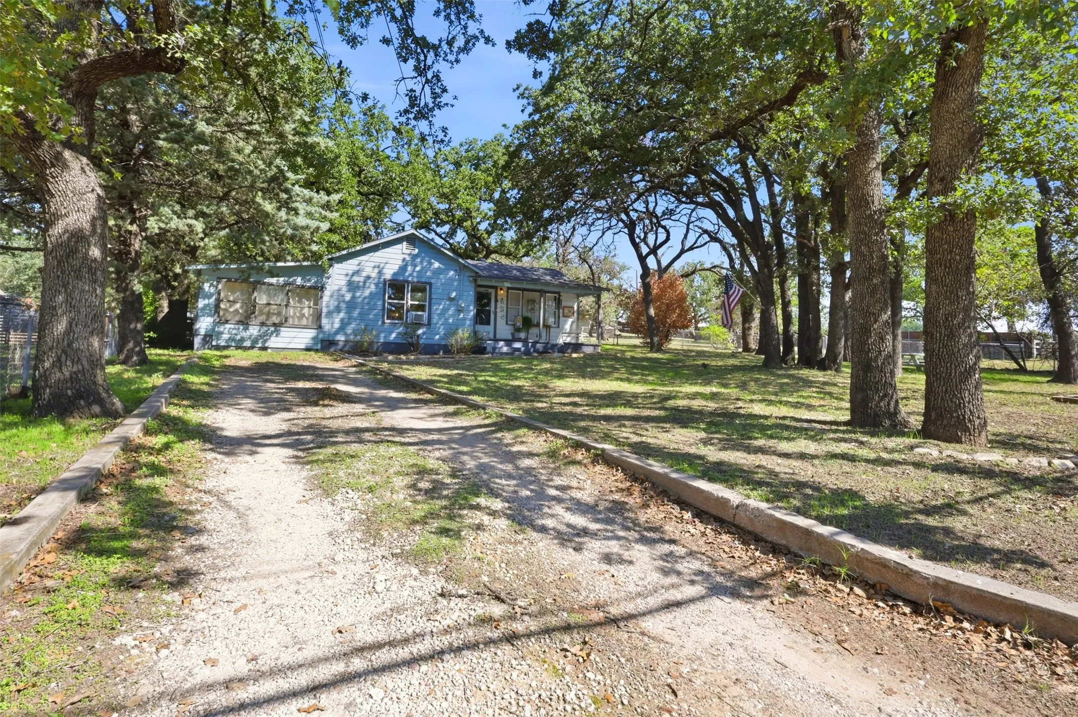 Gravel driveway leading to a light blue exterior residence with a white porch railing, set among mature trees