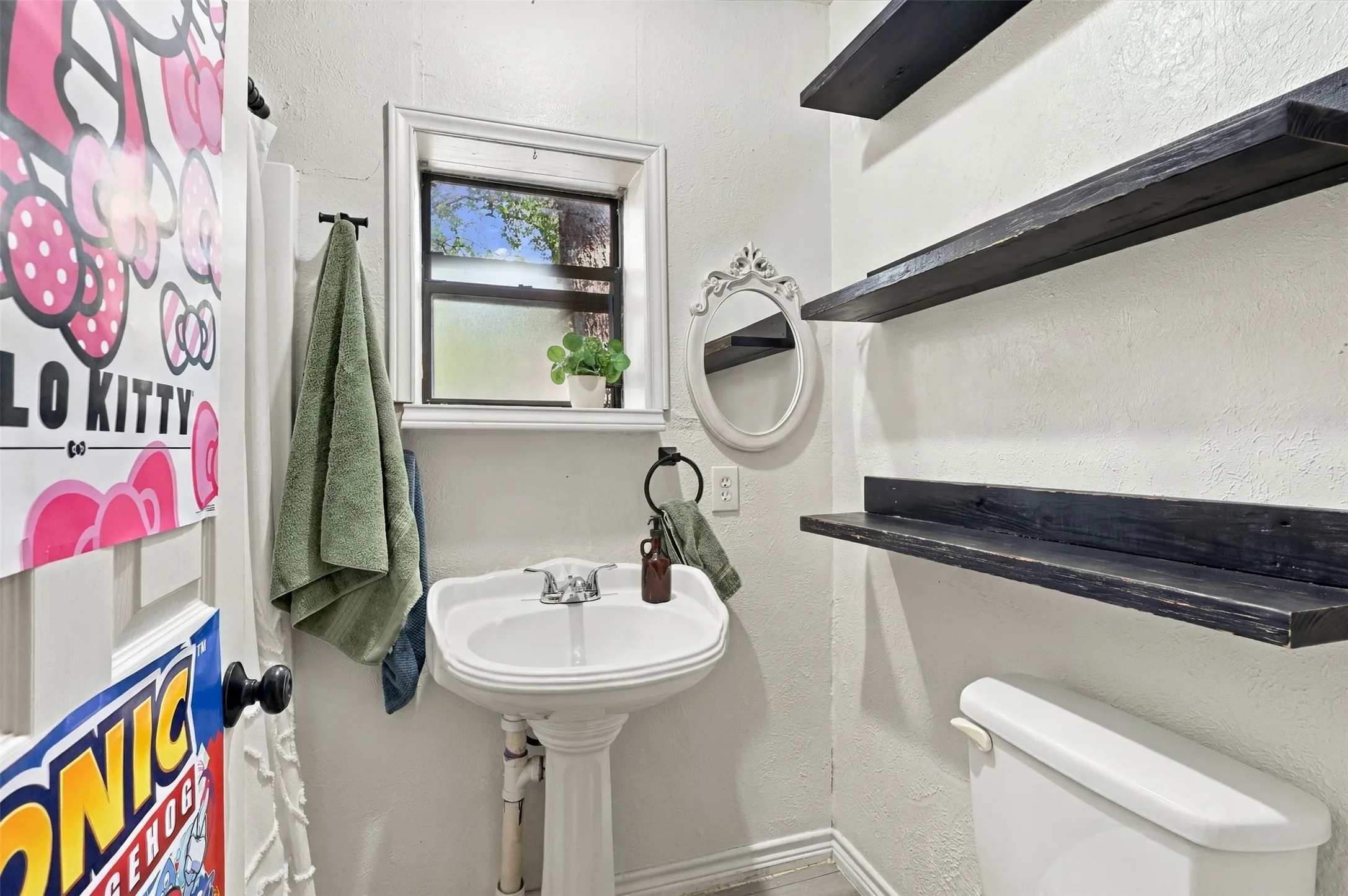 Pedestal sink with chrome fixtures, three dark wood-finish floating shelves, white toilet, and a window with trim