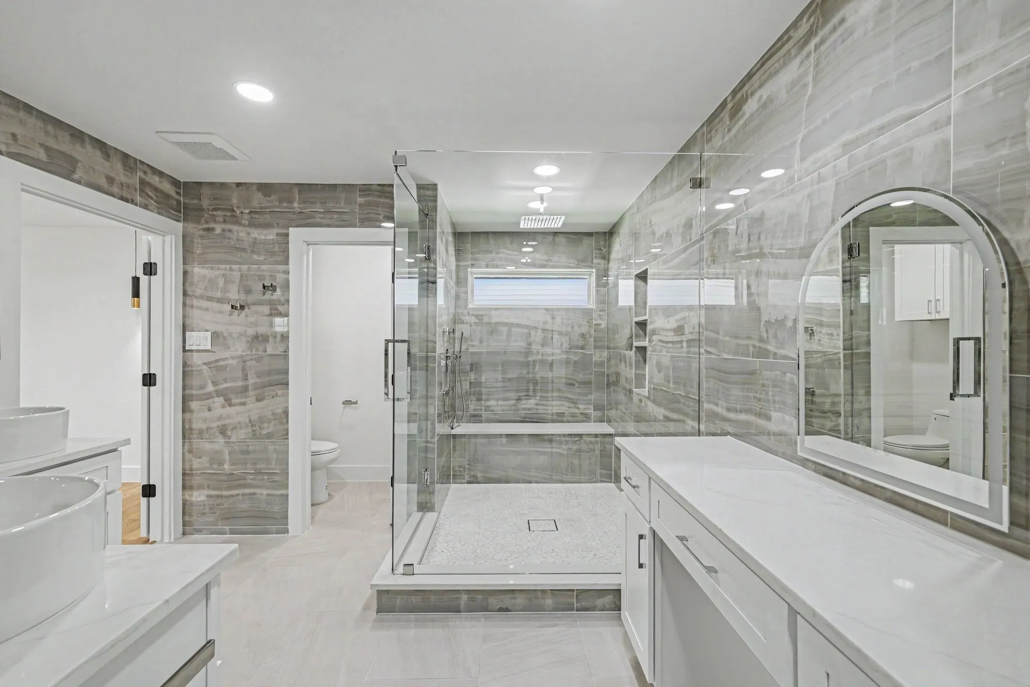Spacious bathroom with dual vanity sinks, extensive white cabinetry, large walk-in shower with glass enclosure, white countertop, and gray tiled walls