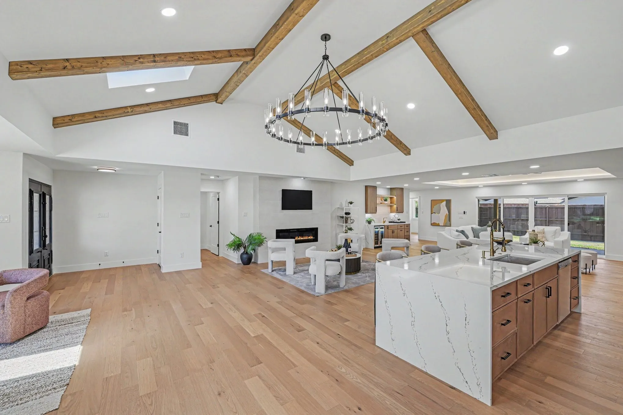 Vaulted ceiling with exposed wood beams, wood-finish flooring, central kitchen island with integrated sink, built-in fireplace, and sliding glass doors