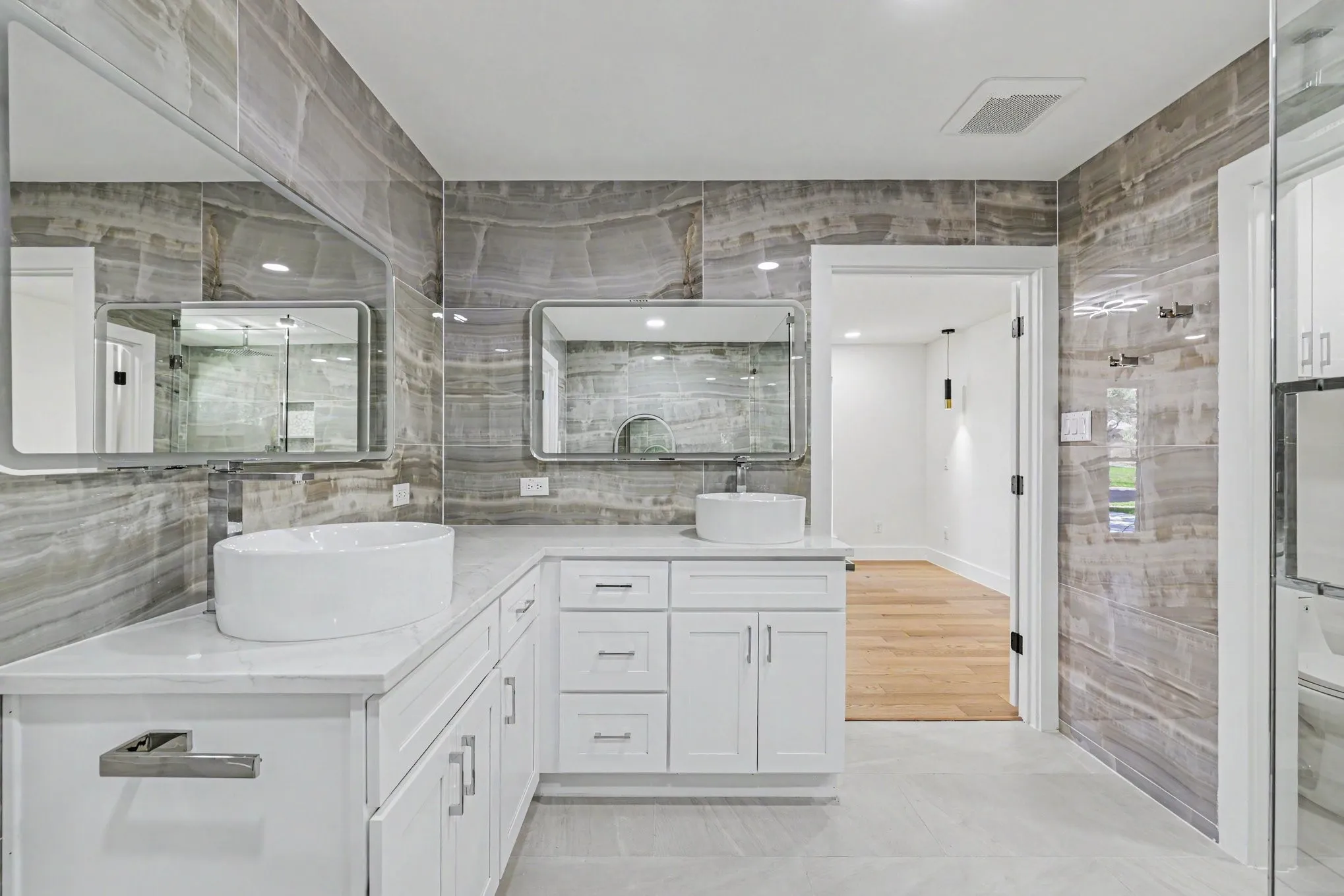 Dual vessel sinks with individual mirrors, custom white cabinetry, and stone-look wall tiling with coordinated flooring