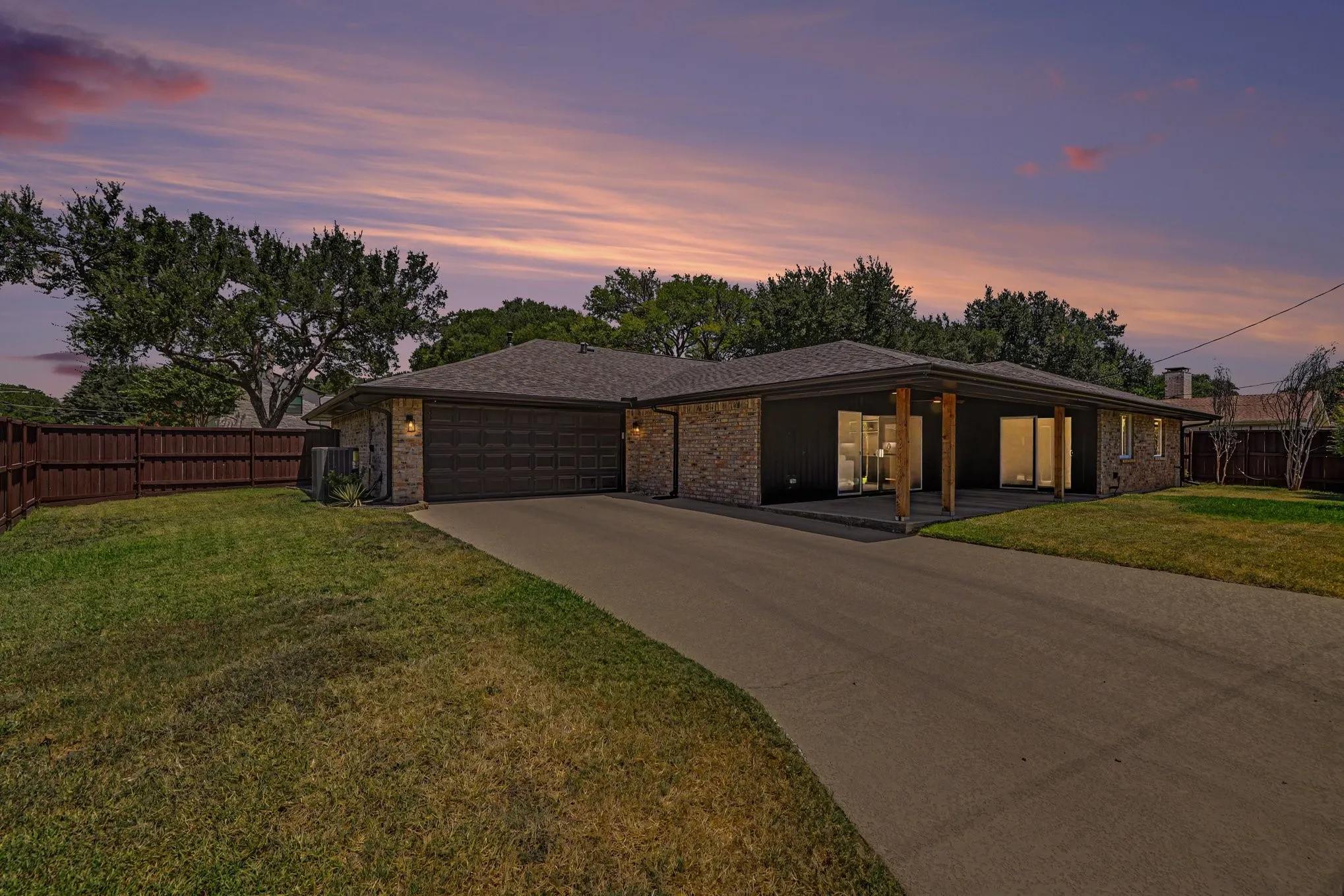 Brick facade with dark siding, covered patio with wooden columns, attached garage, paved driveway, and mature landscaping