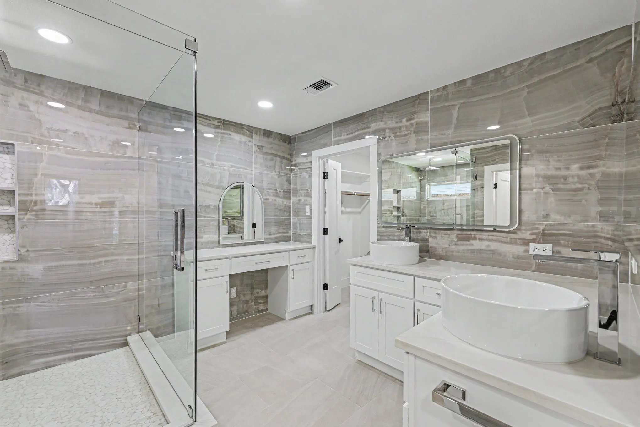 Spacious bathroom featuring a frameless glass shower enclosure, dual vessel sinks, extensive vanity cabinetry, a dedicated dressing vanity, and floor-to-ceiling stone-look wall tiling