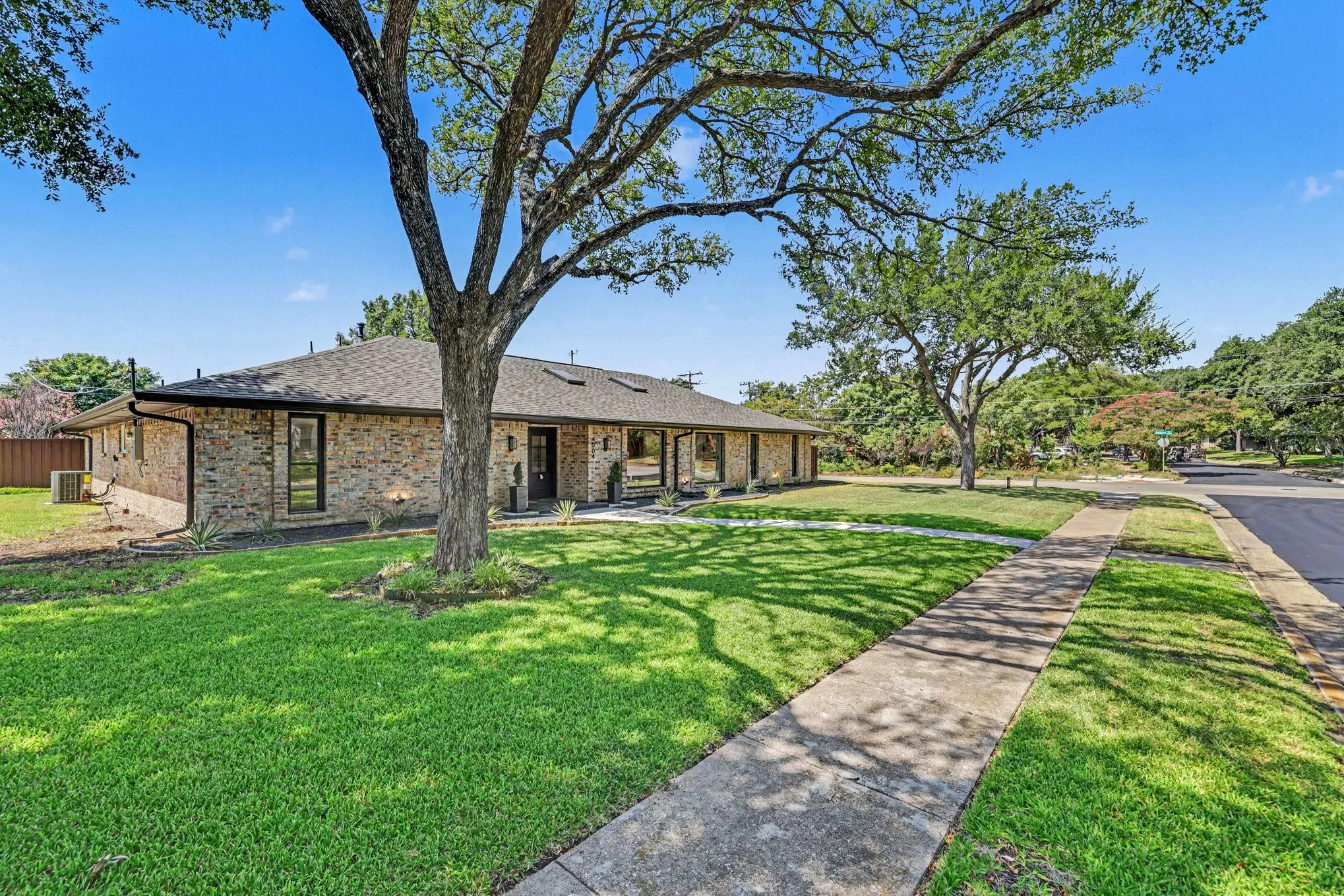 Brick exterior with a dark shingle roof, mature trees, concrete sidewalk, and manicured lawn