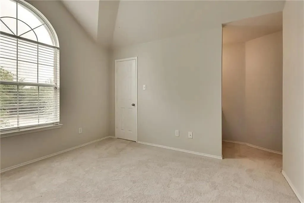 Arched window with blinds, vaulted ceiling, neutral wall paint, light-colored carpeting, and a white paneled door