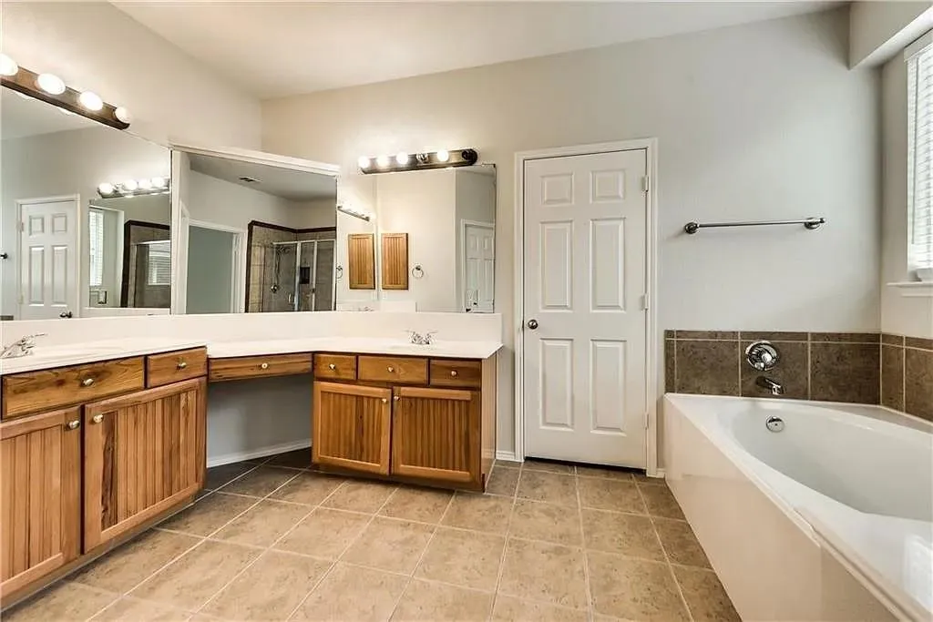 Dual vanities with integrated sinks, wood-finish cabinetry, ceramic tile flooring, built-in bathtub, and horizontal blinds