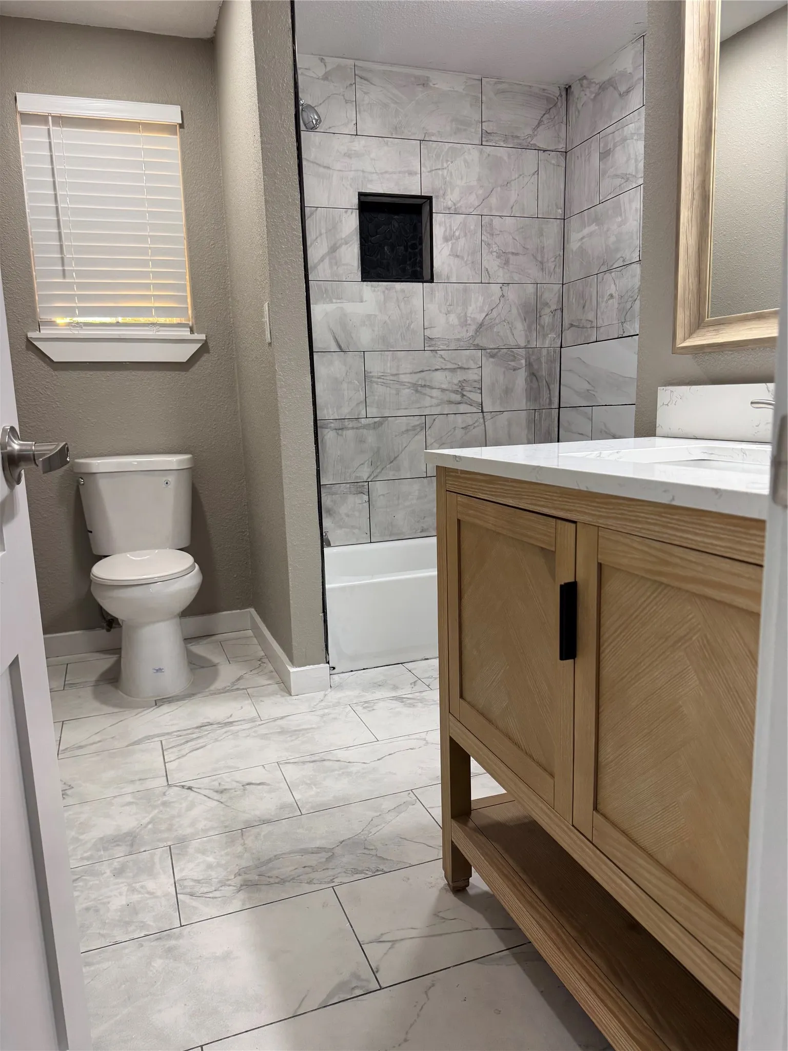 Bathroom featuring a light wood-finish vanity with black hardware, white countertop with an integrated sink, and large format floor tile
