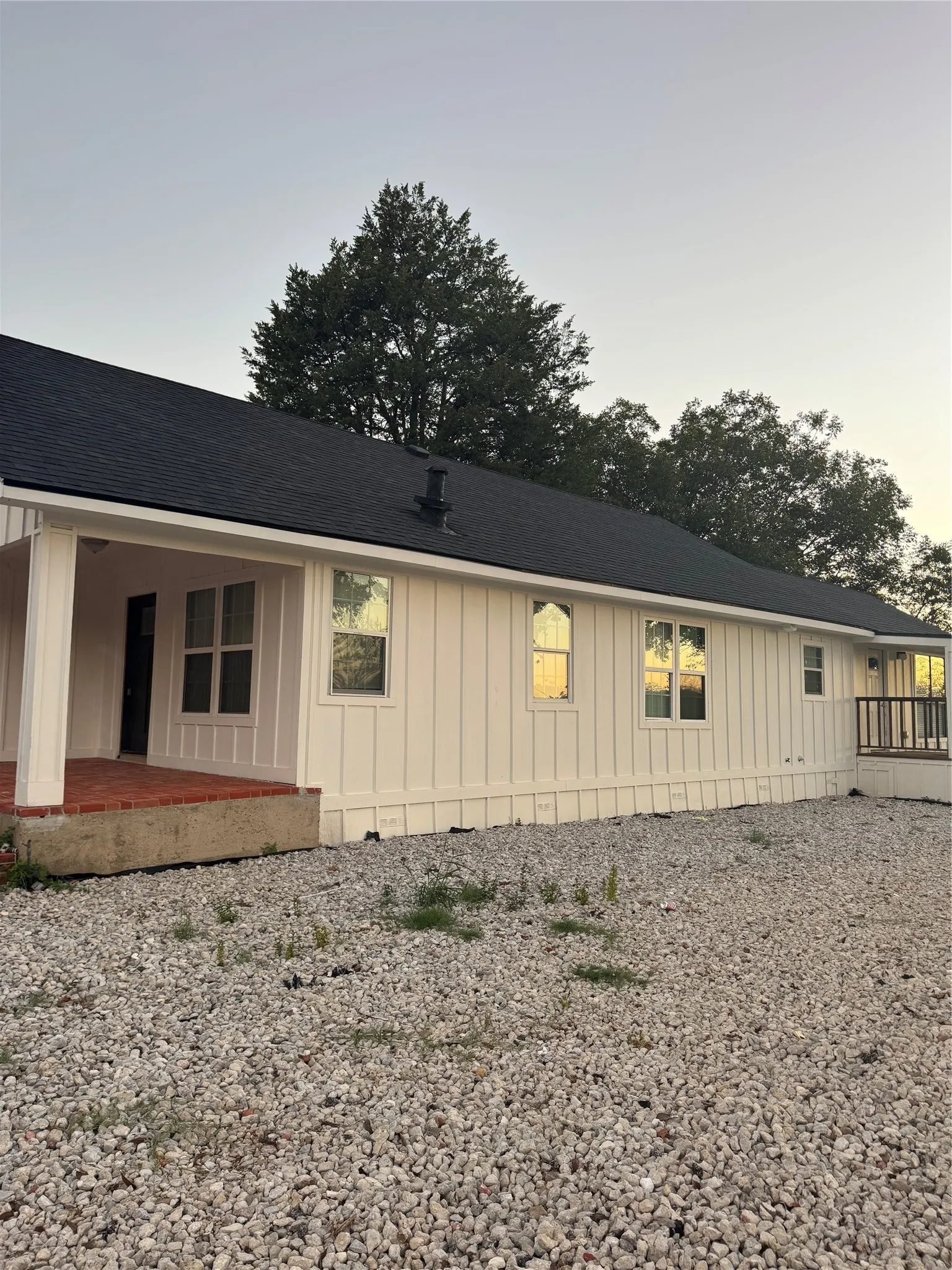 White vertical board and batten siding, dark shingle roof, covered entry porch with brick base, multiple windows, and gravel ground cover