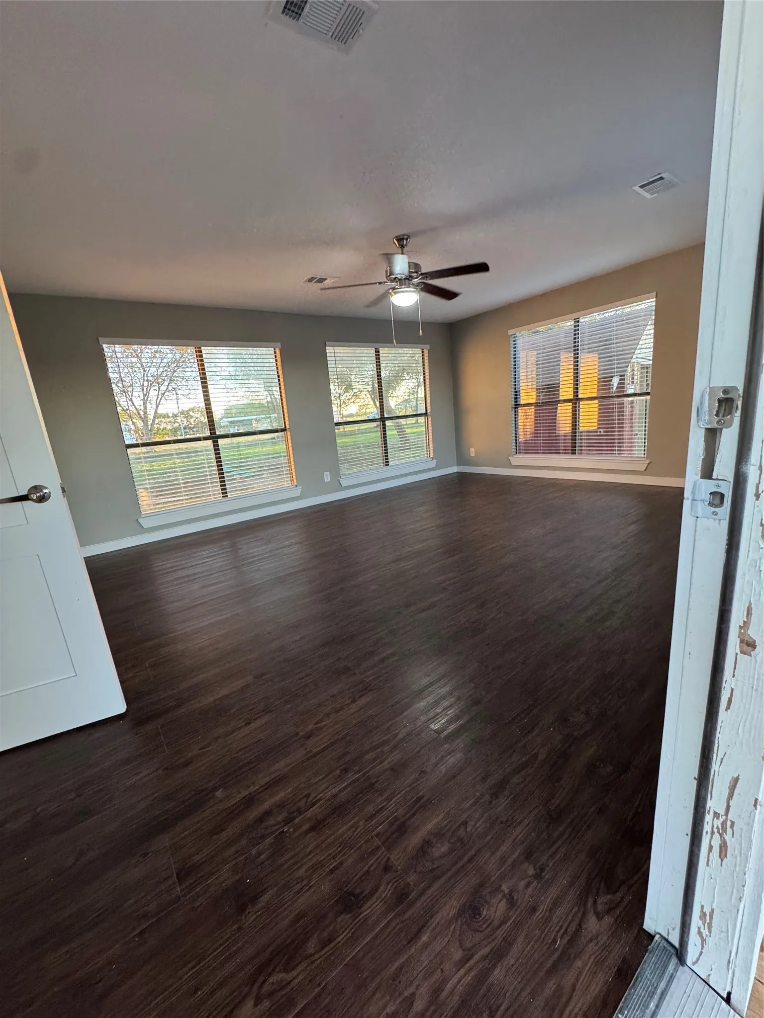Spacious room featuring dark wood-finish flooring, multiple windows with blinds, and a ceiling fan with integrated lighting