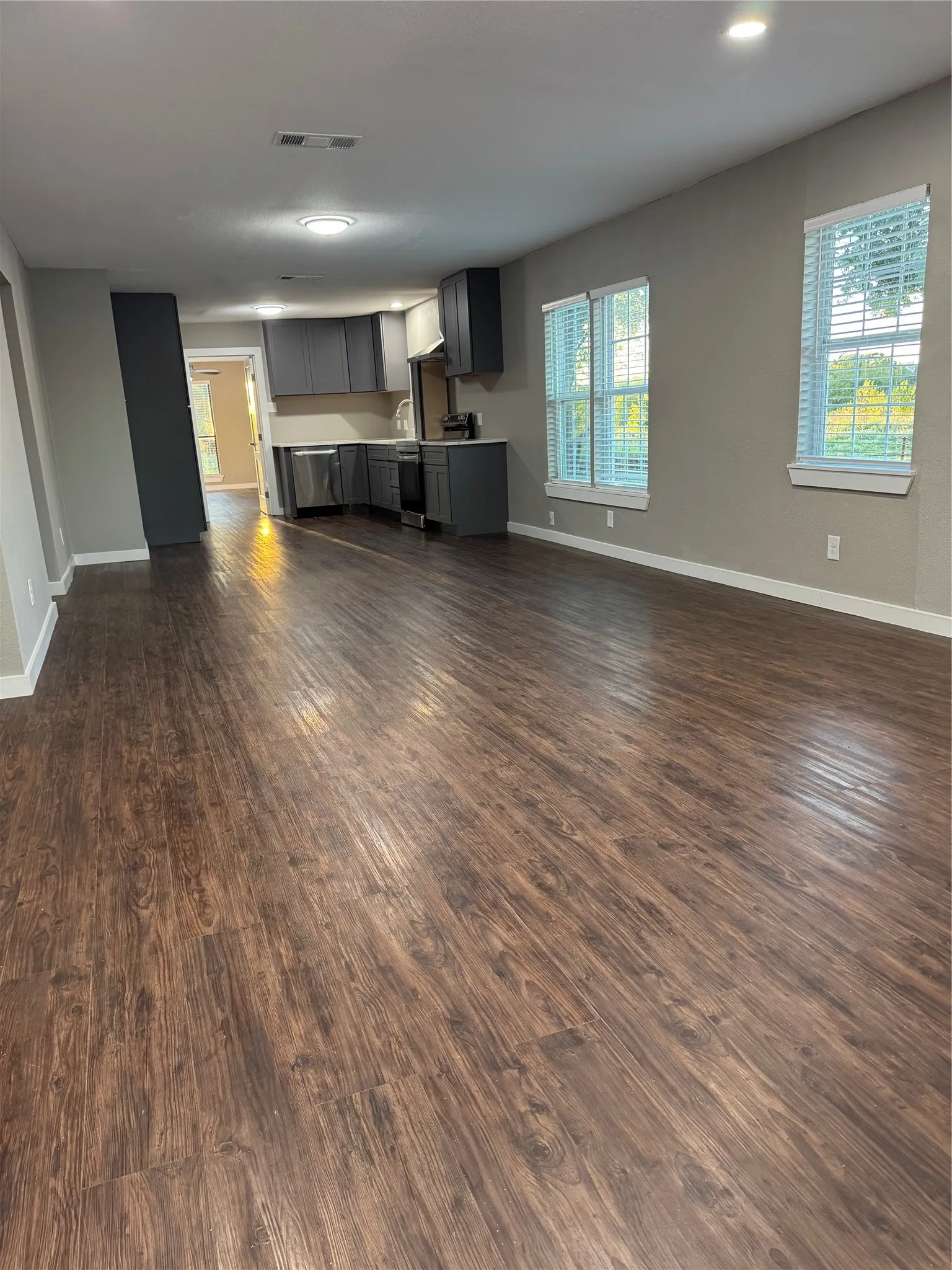 Wood-finish flooring throughout, recessed ceiling lights, multiple windows, and a kitchen featuring dark cabinetry and stainless steel appliances