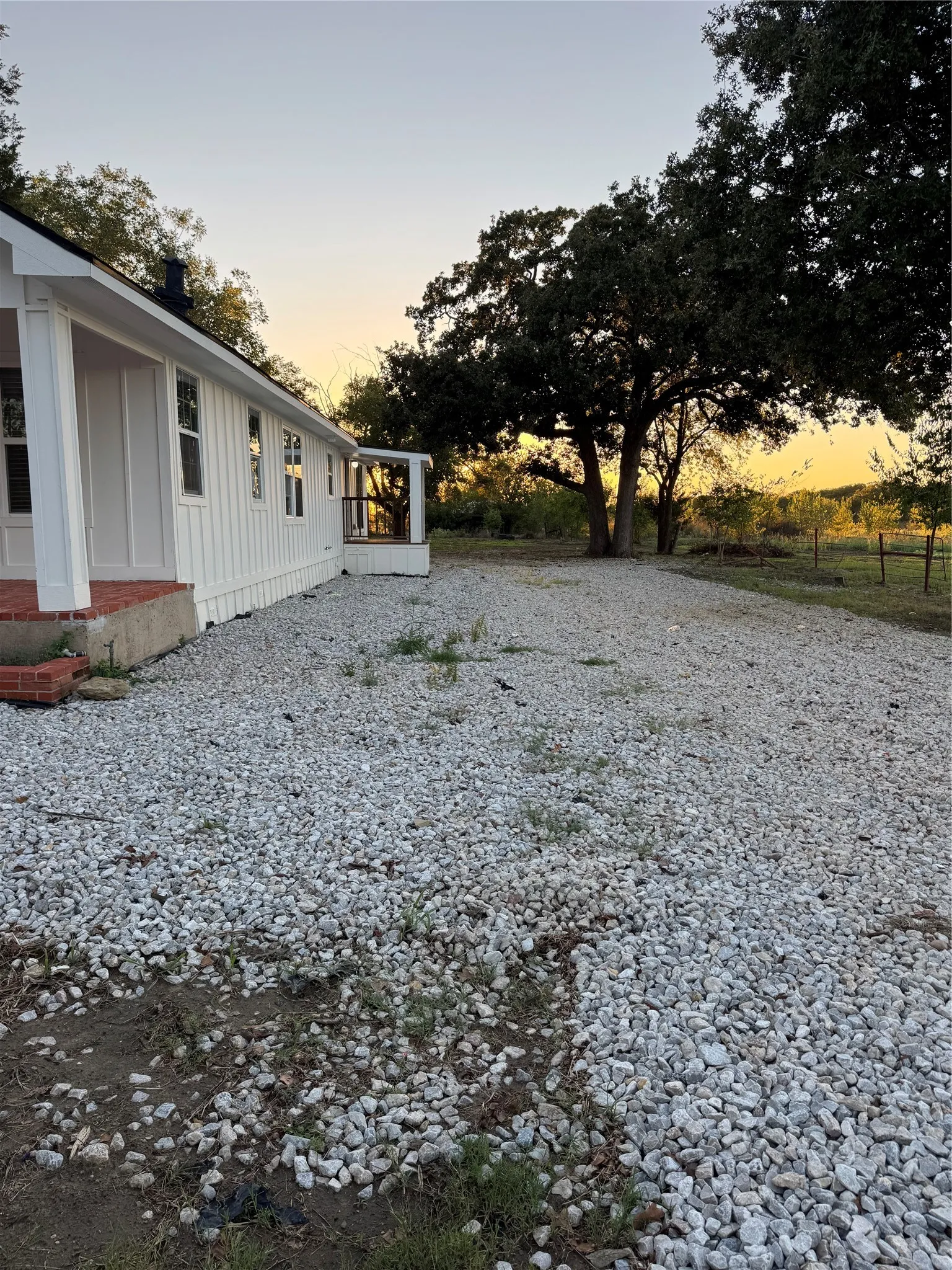 White exterior with vertical siding, covered porch with brick foundation, gravel ground cover, and mature trees