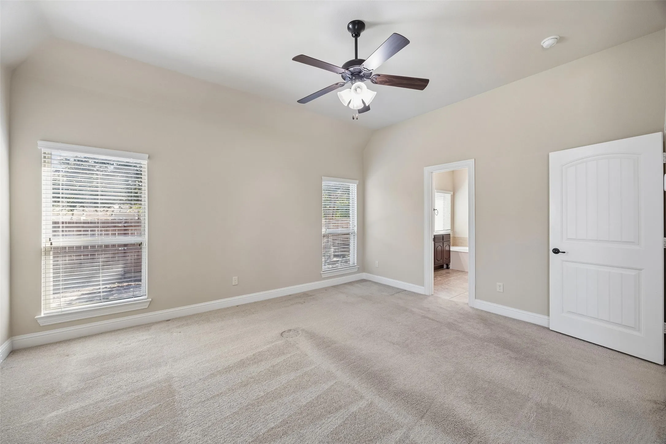 Vaulted ceiling with a ceiling fan light fixture, two windows with blinds, a white paneled door, and neutral-toned carpeting