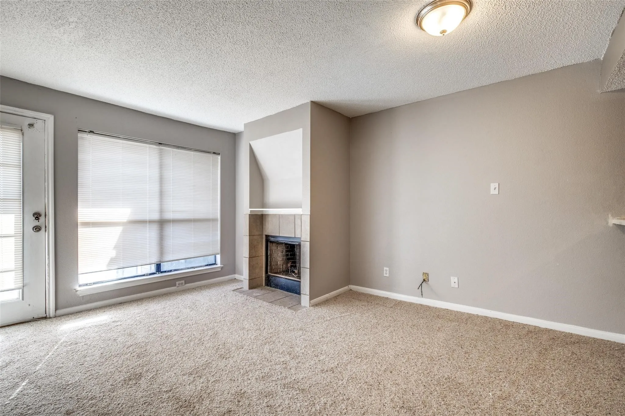 Neutral tone wall paint, textured ceiling, built-in fireplace with tile surround, window with blinds, and light carpet flooring