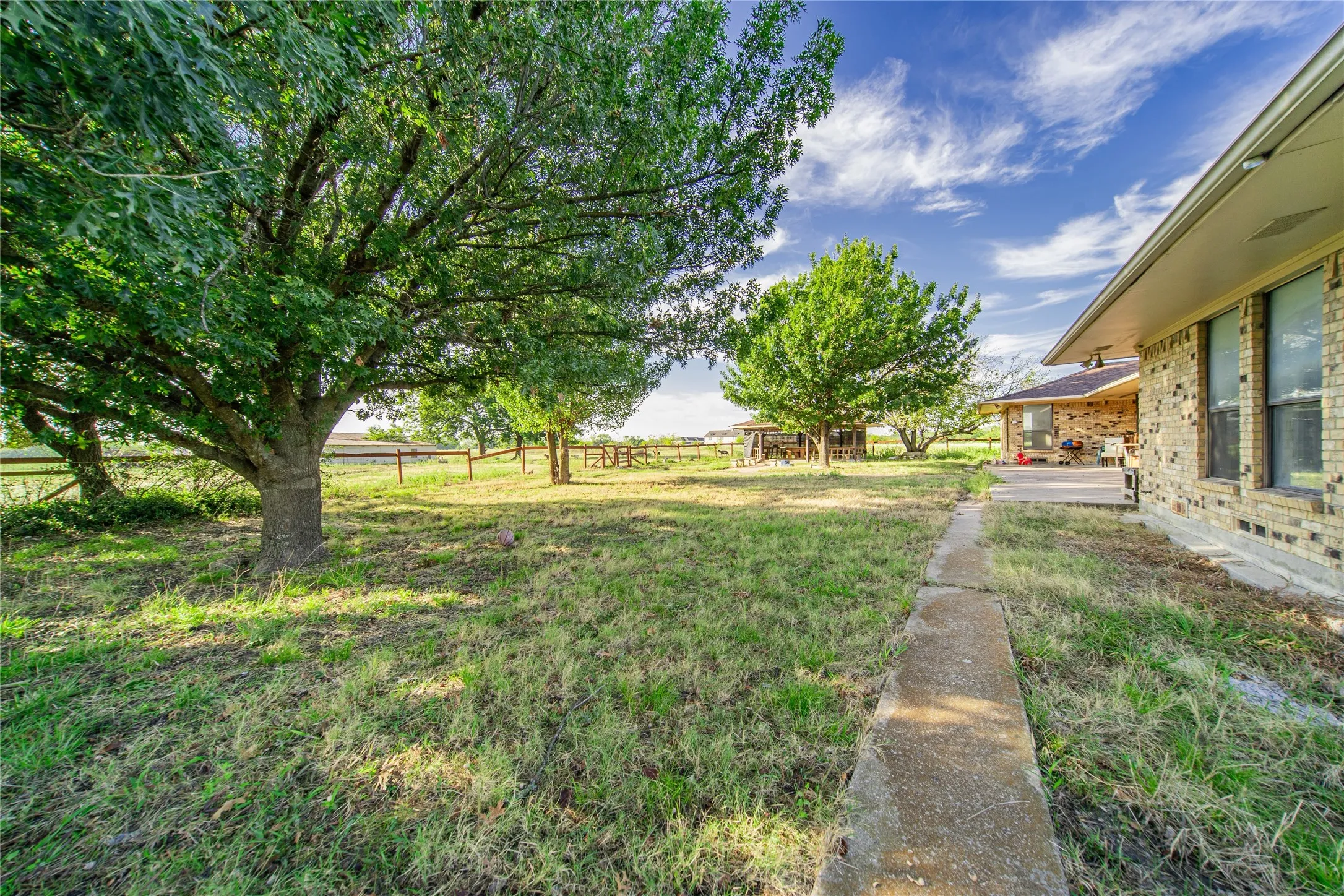 Expansive grassy area with mature trees and a concrete pathway leading to a covered patio