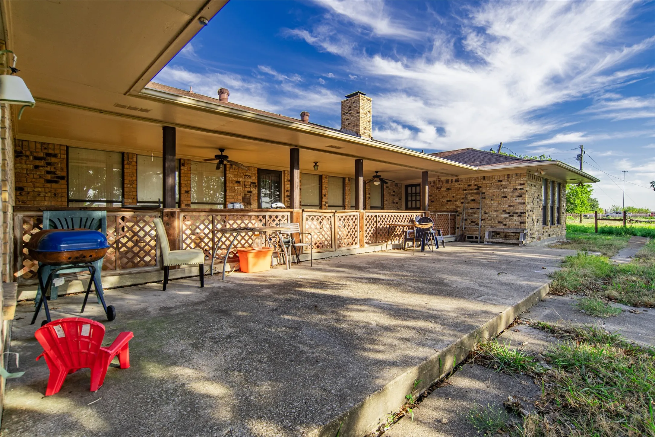 Expansive concrete patio with a covered porch featuring multiple support columns, a wood-lattice railing, and a ceiling fan