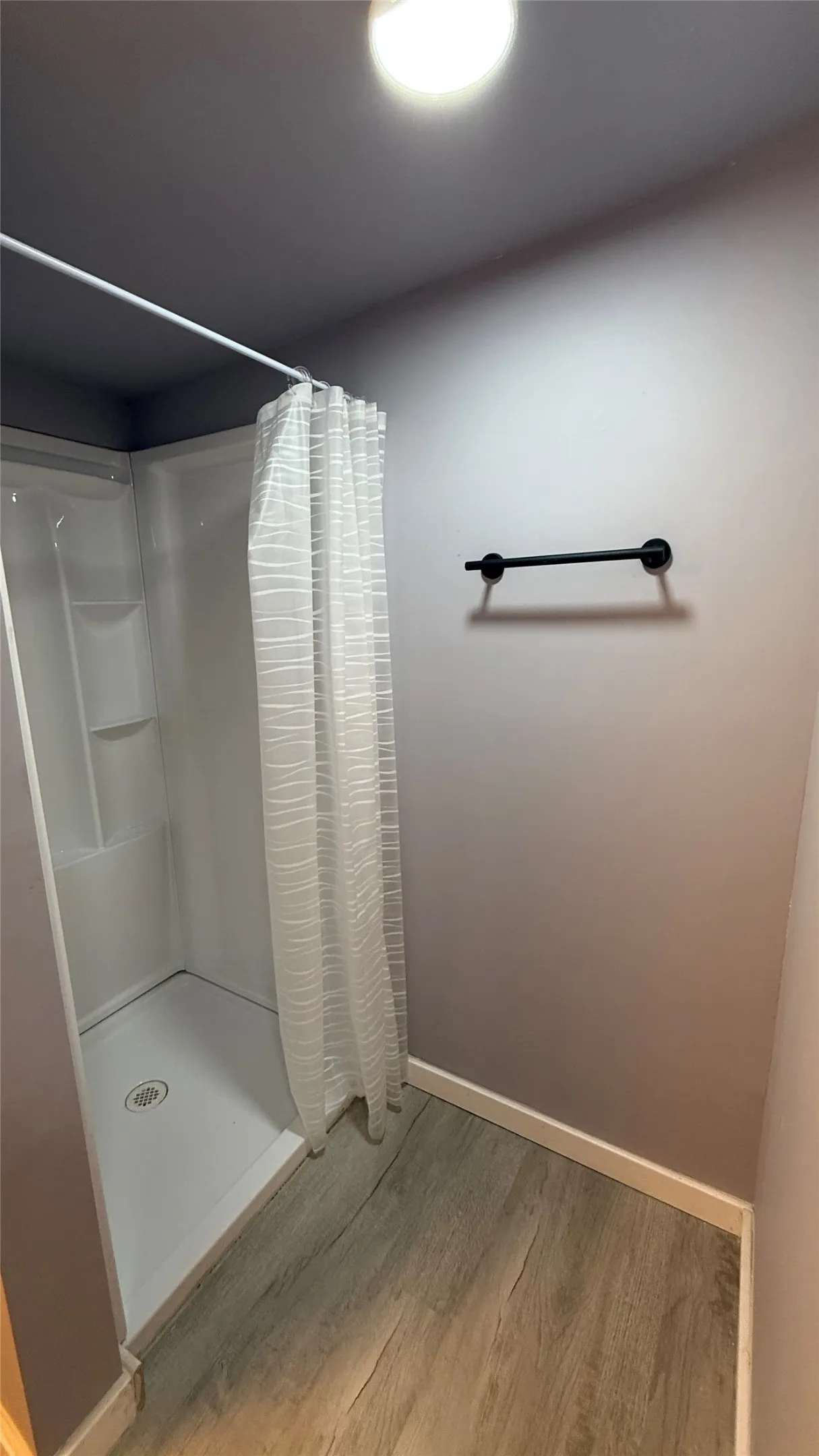 Shower stall with white surround, wood-finish flooring, and a matte black towel bar