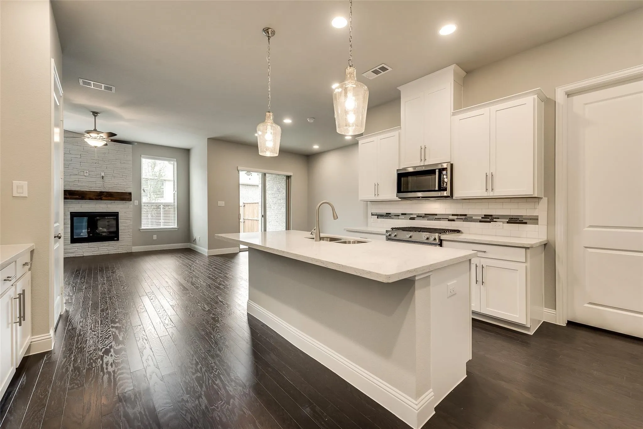 Kitchen island with a built-in sink and pendant lighting, white cabinetry, dark wood-finish flooring, a contemporary fireplace with a wood mantel, and sliding glass doors