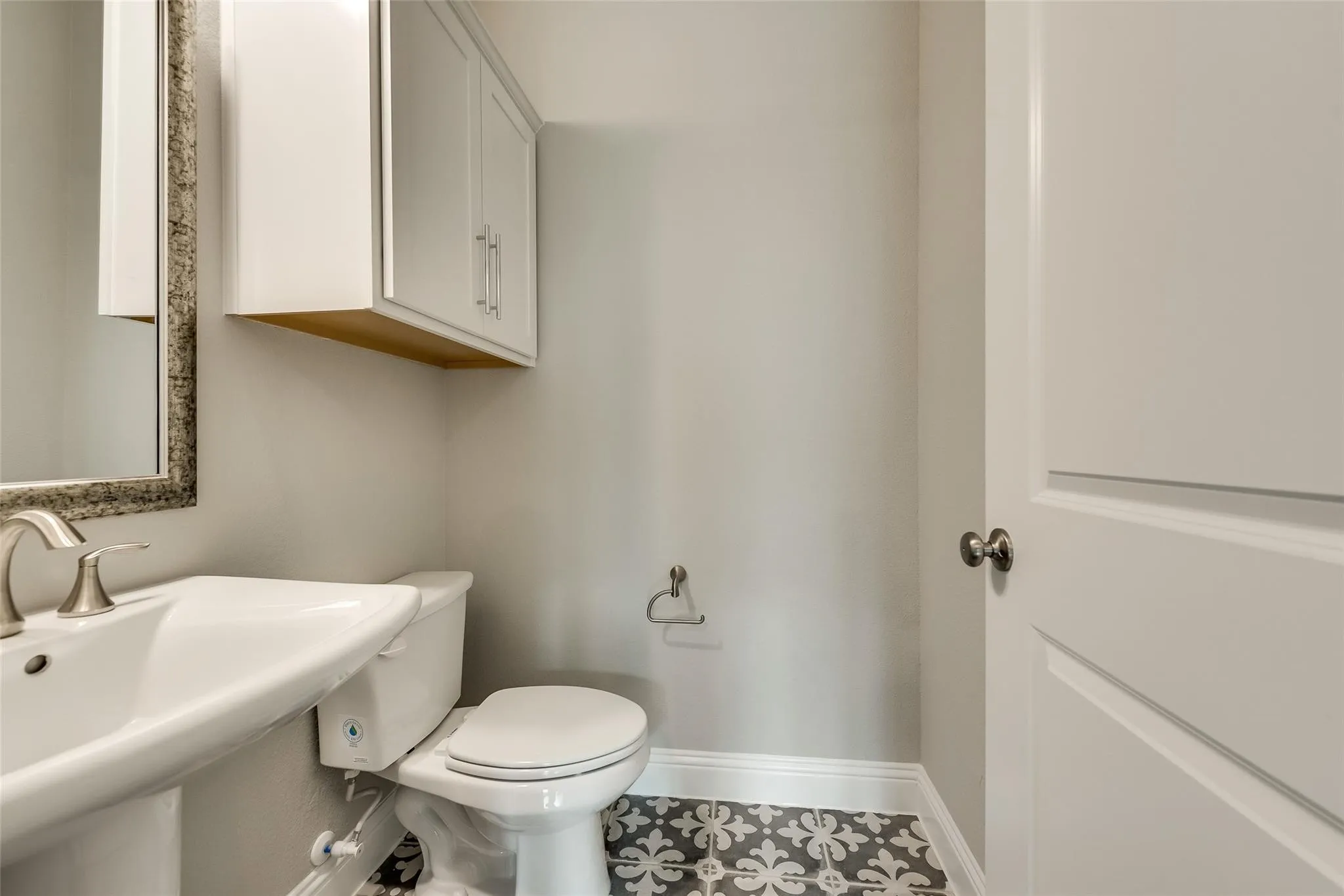 Pedestal sink with a brushed nickel faucet, framed mirror, wall-mounted cabinetry, and decorative floor tiling