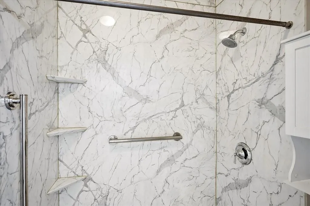 White marble-patterned shower walls, polished chrome shower fixtures, corner storage shelves, and a built-in grab bar