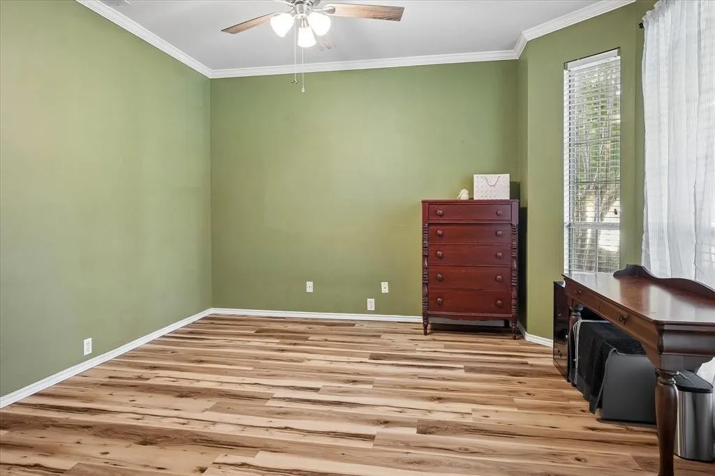 Wood-finish flooring, ceiling fan with integrated lighting, crown molding, and a corner window with horizontal blinds