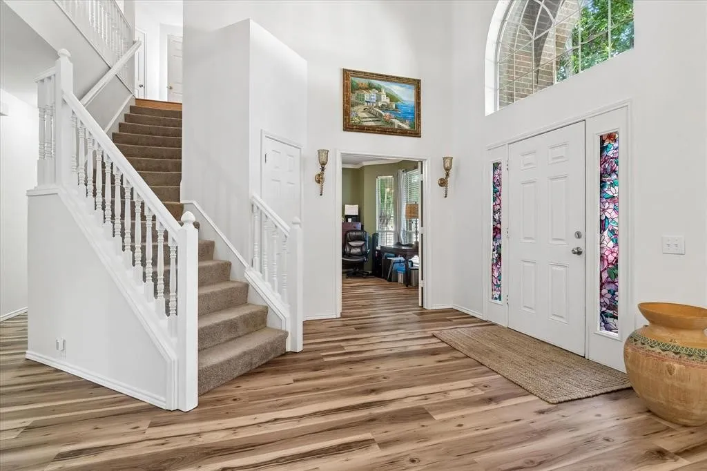 Two-story foyer with wood-finish flooring, white staircase with carpeted steps, and a double-door entry with decorative glass side panels