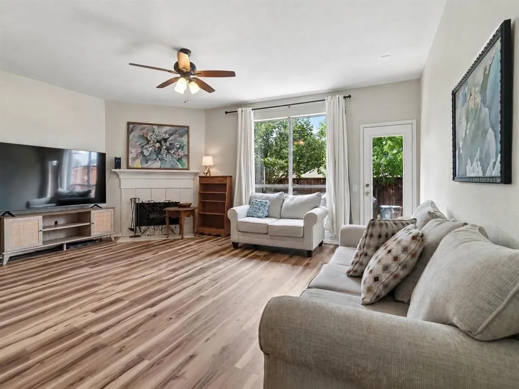 Wood-finish flooring, fireplace with white mantel, ceiling fan with integrated lighting, glass-paneled exterior door, and large window