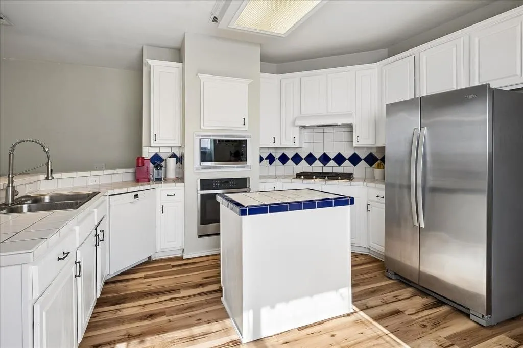 Kitchen featuring white cabinetry, tile countertops, wood-finish flooring, built-in stainless steel appliances, and a central island with tile trim