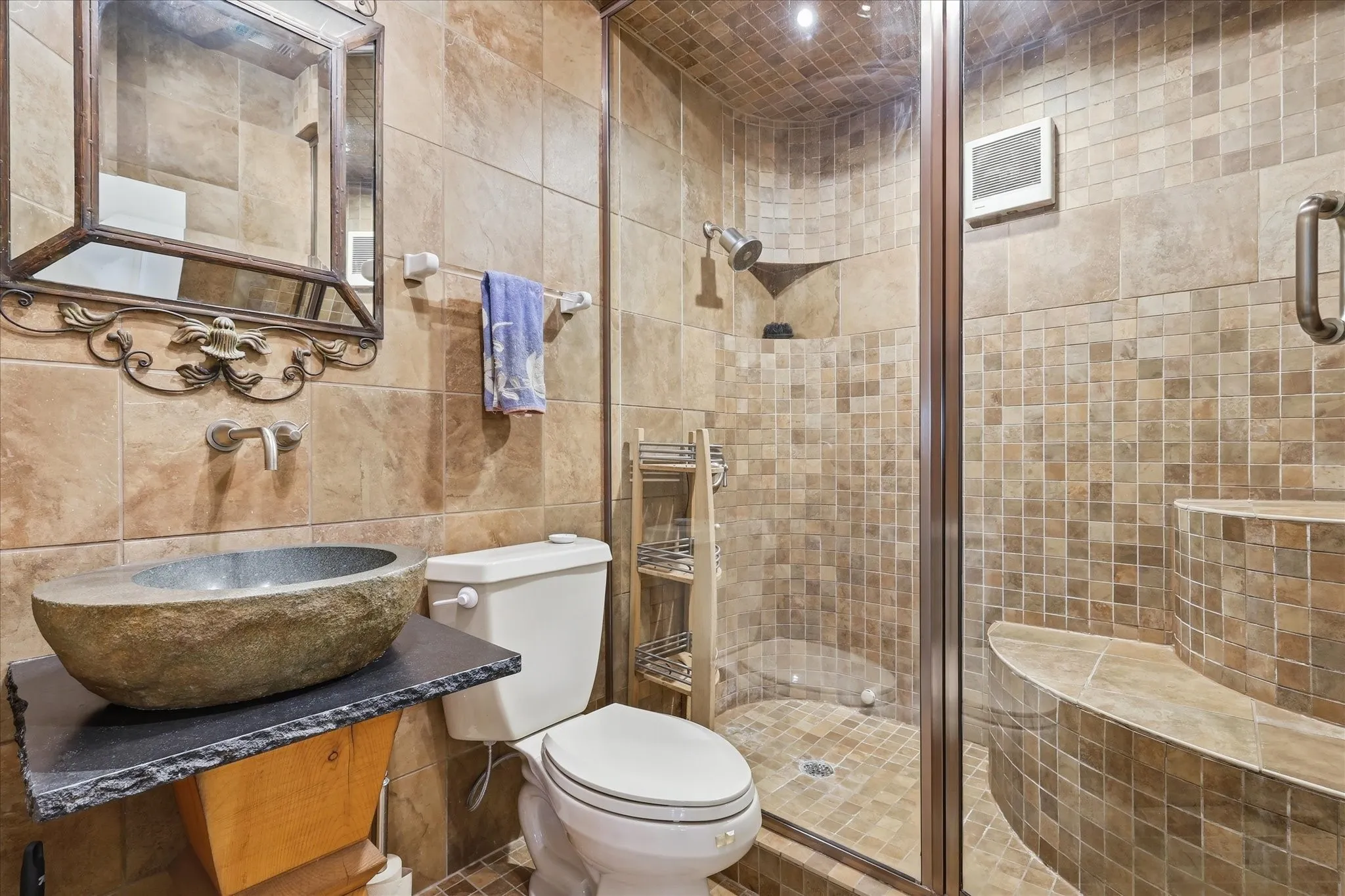 Stone vessel sink with a black slate countertop, tiled shower enclosure with glass door, fixed shower head, decorative mirror, and built-in shower shelving
