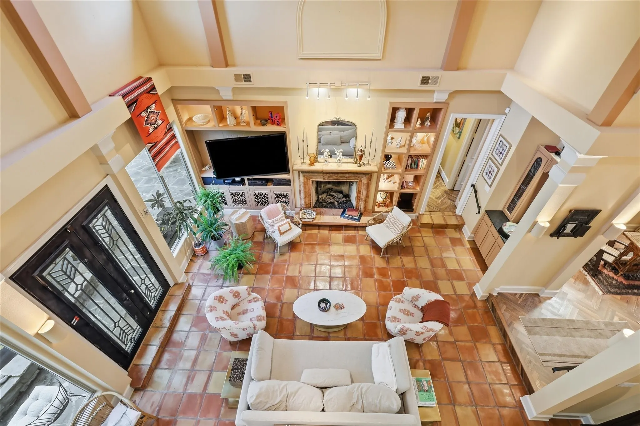 Vaulted ceiling with exposed beams, terracotta tile flooring, built-in shelving, stone fireplace surround, and decorative glass double entry doors