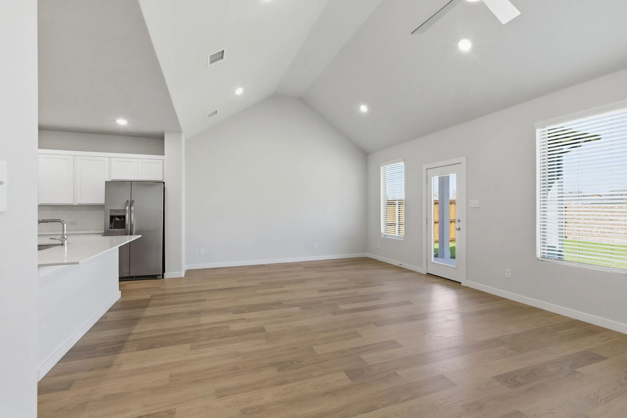 Vaulted ceiling with recessed lighting, wood-finish flooring, white kitchen cabinetry, stainless steel refrigerator, and a kitchen island with sink