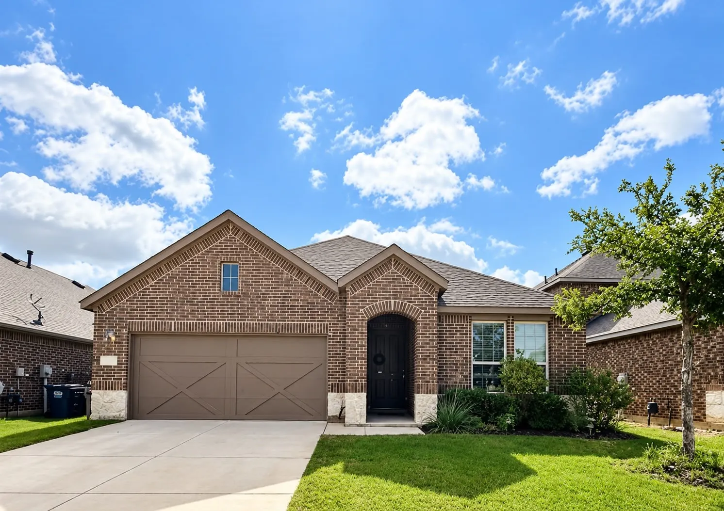 Brick exterior with stone accents, arched entryway, attached garage with decorative doors, and mature landscaping