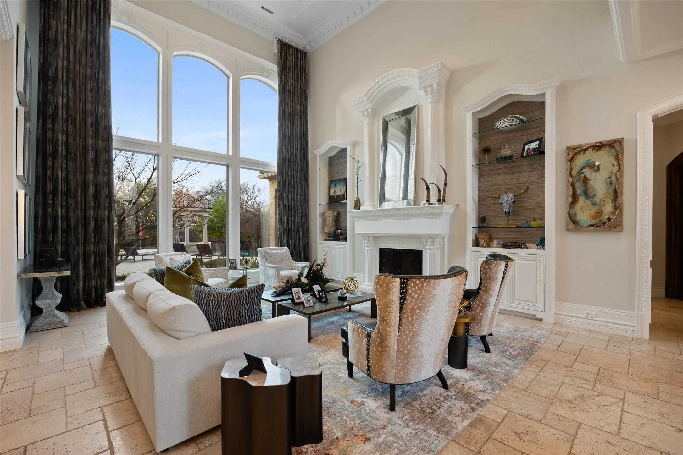 Soaring ceilings with decorative crown molding, full-height arched windows, a stone fireplace with ornate mantel, and built-in shelving with glass fronts