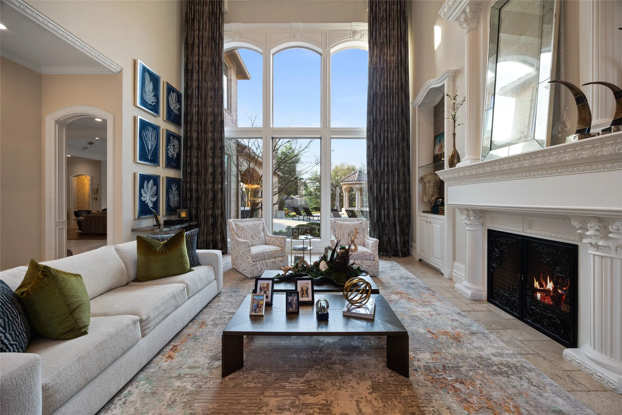 Two-story living area with full-height windows, ornate fireplace with custom mantel, built-in cabinetry, arched doorways, and tile flooring