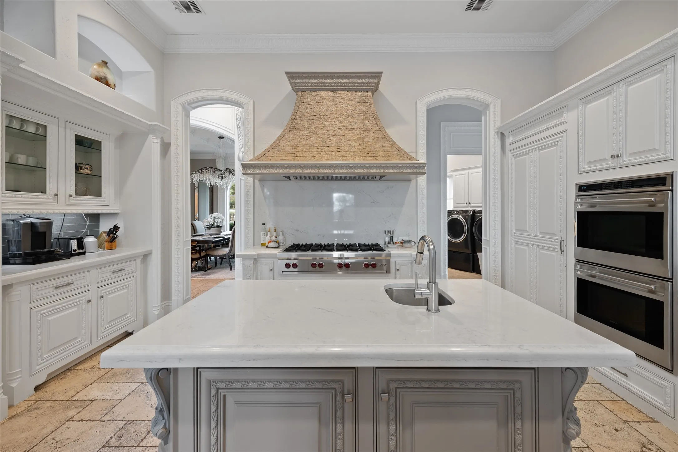 Central island with integrated sink, stone countertops, custom range hood, built-in double ovens, and decorative cabinetry