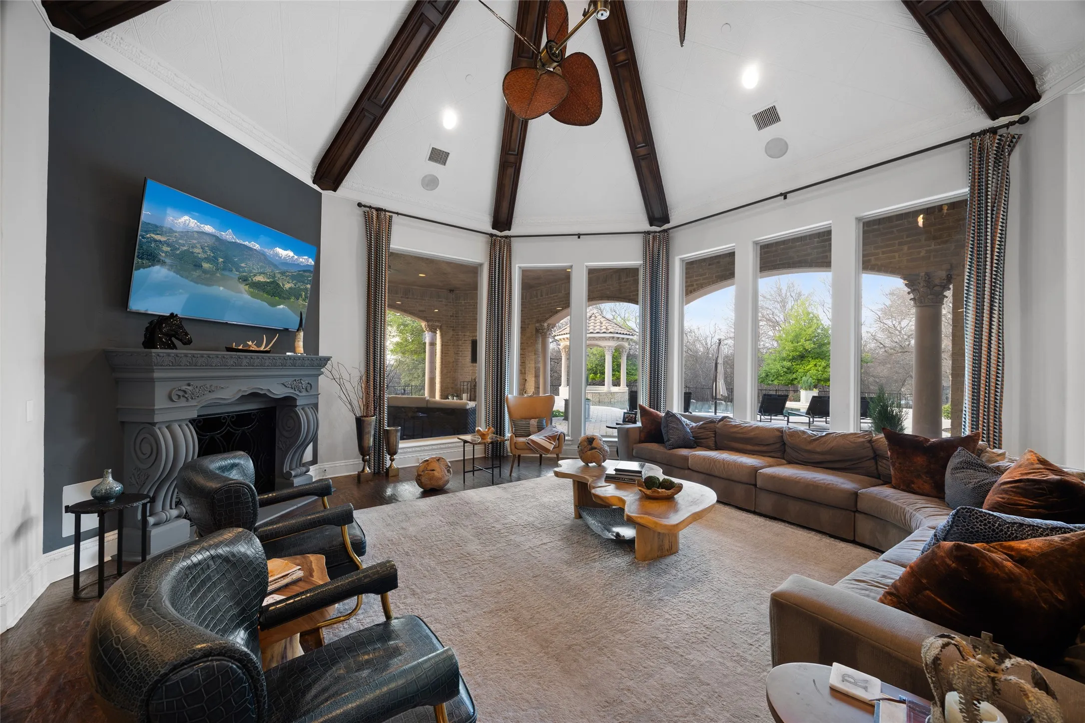 Vaulted ceiling with exposed wood beams, ornate fireplace with mantel, dark wood-finish flooring, and multiple large windows