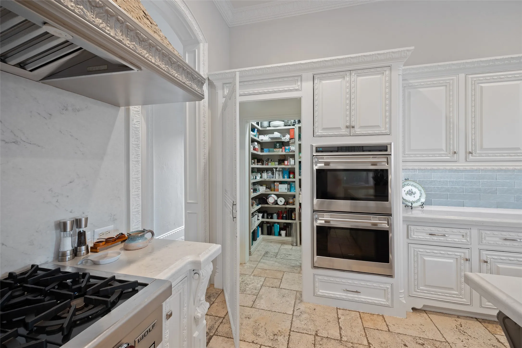 White cabinetry with decorative molding, stainless steel double ovens, tile backsplash, and a walk-in pantry