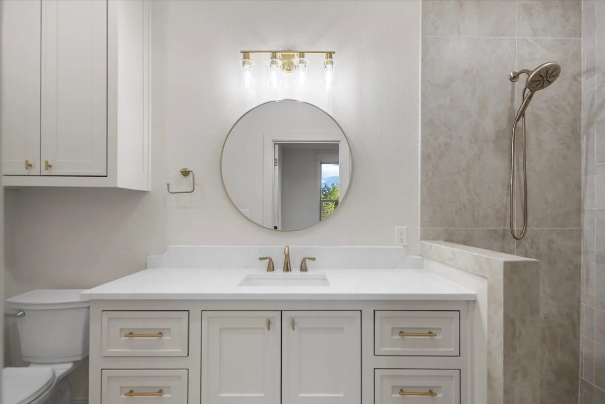 White shaker cabinetry with gold-finish hardware, a single vanity with an undermount sink, a round mirror, and a gold-finish vanity light fixture