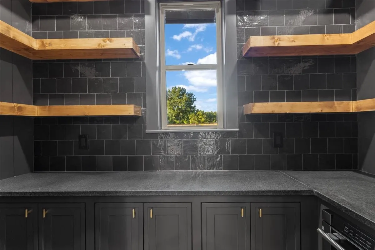 Built-in cabinetry with gold-finish hardware, dark stone countertops, dark tiled backsplash, and natural wood floating shelves