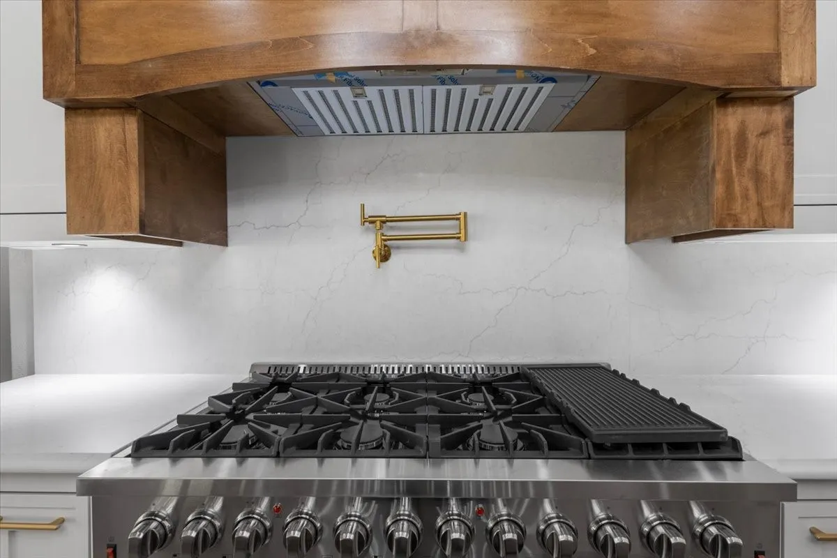 Professional-grade range with a built-in griddle, wood-finish range hood, white countertop, and a brass pot filler