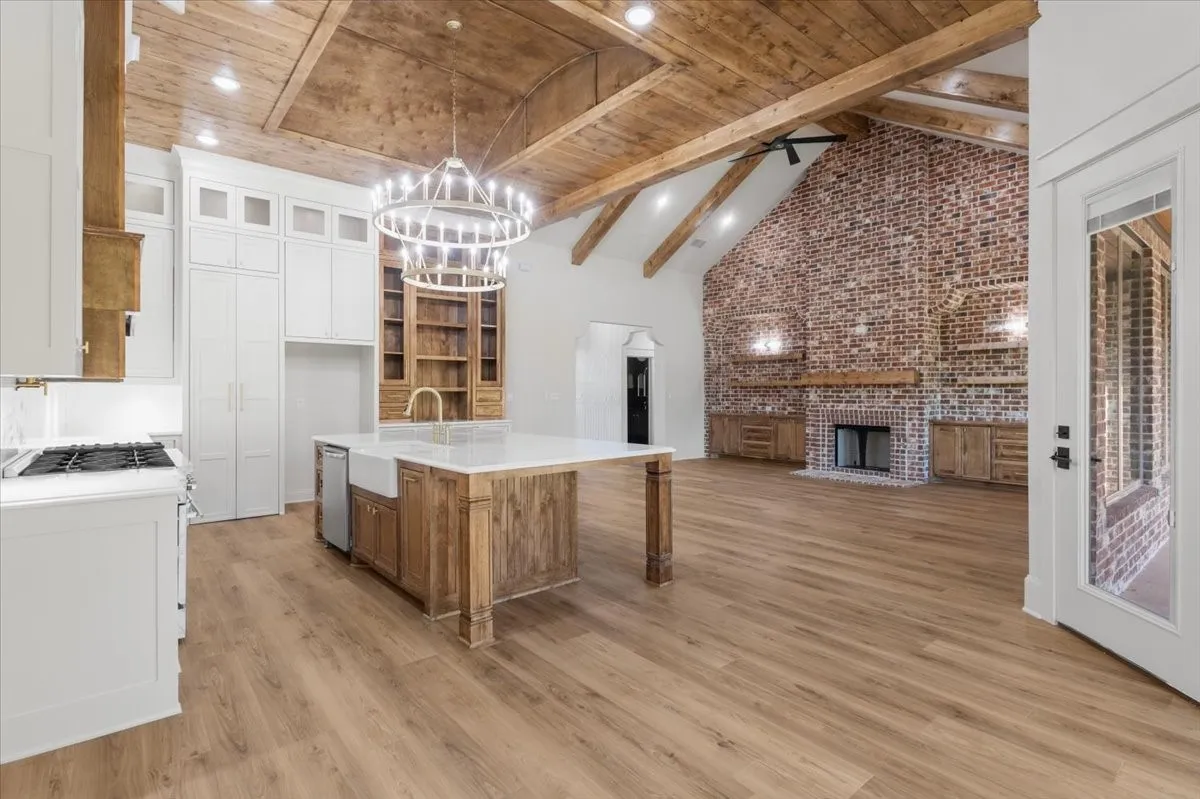 Vaulted wood-beamed ceiling with exposed trusses, wood-finish flooring, brick accent wall with fireplace, white shaker cabinetry, and a kitchen island with a farmhouse sink