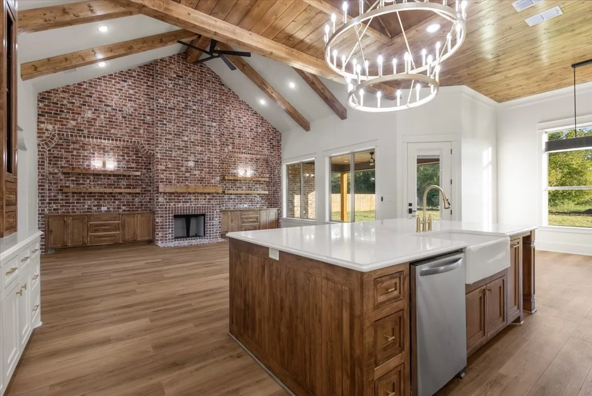 Vaulted ceiling with exposed wood beams, brick accent wall with fireplace, wood-finish flooring, kitchen island with built-in sink and dishwasher, and large glass pane windows