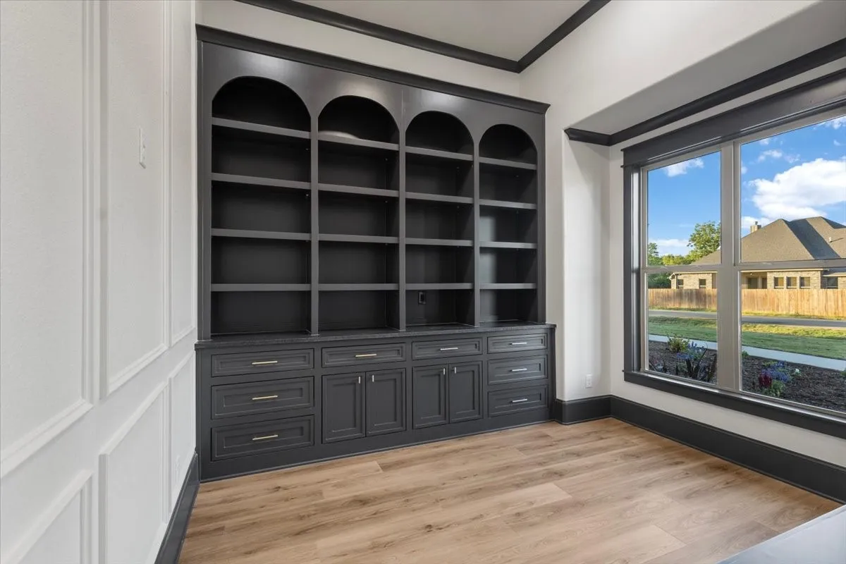 Built-in shelving with arched tops and lower cabinetry, wood-finish flooring, large window with dark trim, wainscoting detail on walls, and dark crown molding