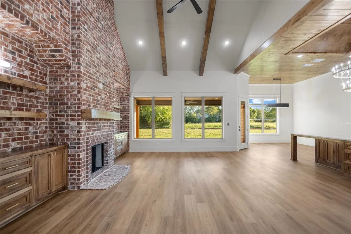 Soaring vaulted ceiling with exposed wood beams, brick fireplace with wood mantle, wood-finish flooring, recessed lighting, and large windows