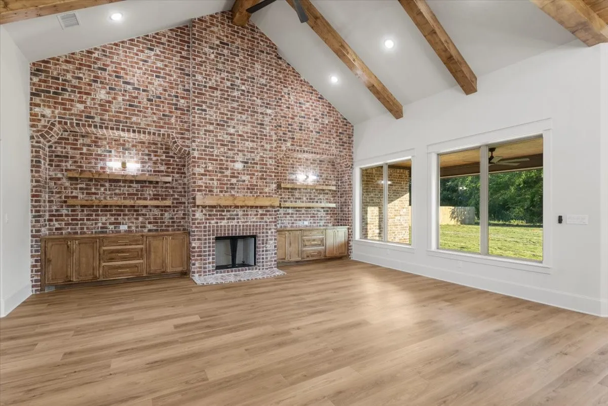 Vaulted ceiling with exposed wood beams, full-height brick accent wall with fireplace, built-in wooden cabinetry, wood-finish flooring, and large windows